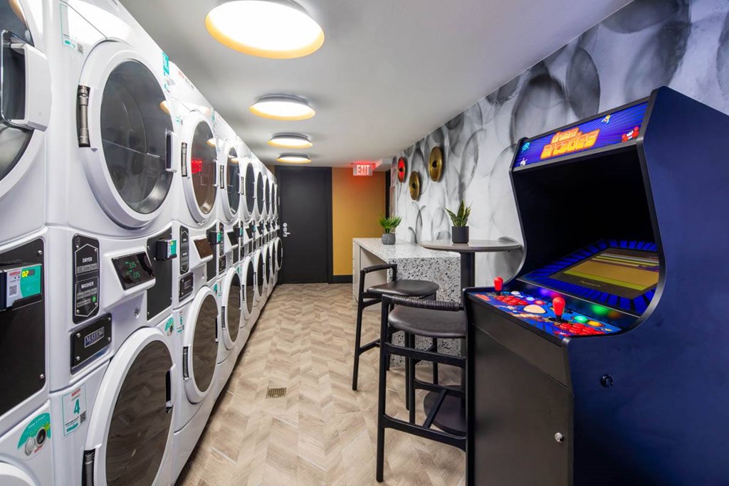 A row of washing machines are lined up in a room with a pinball machine and a bar stool.