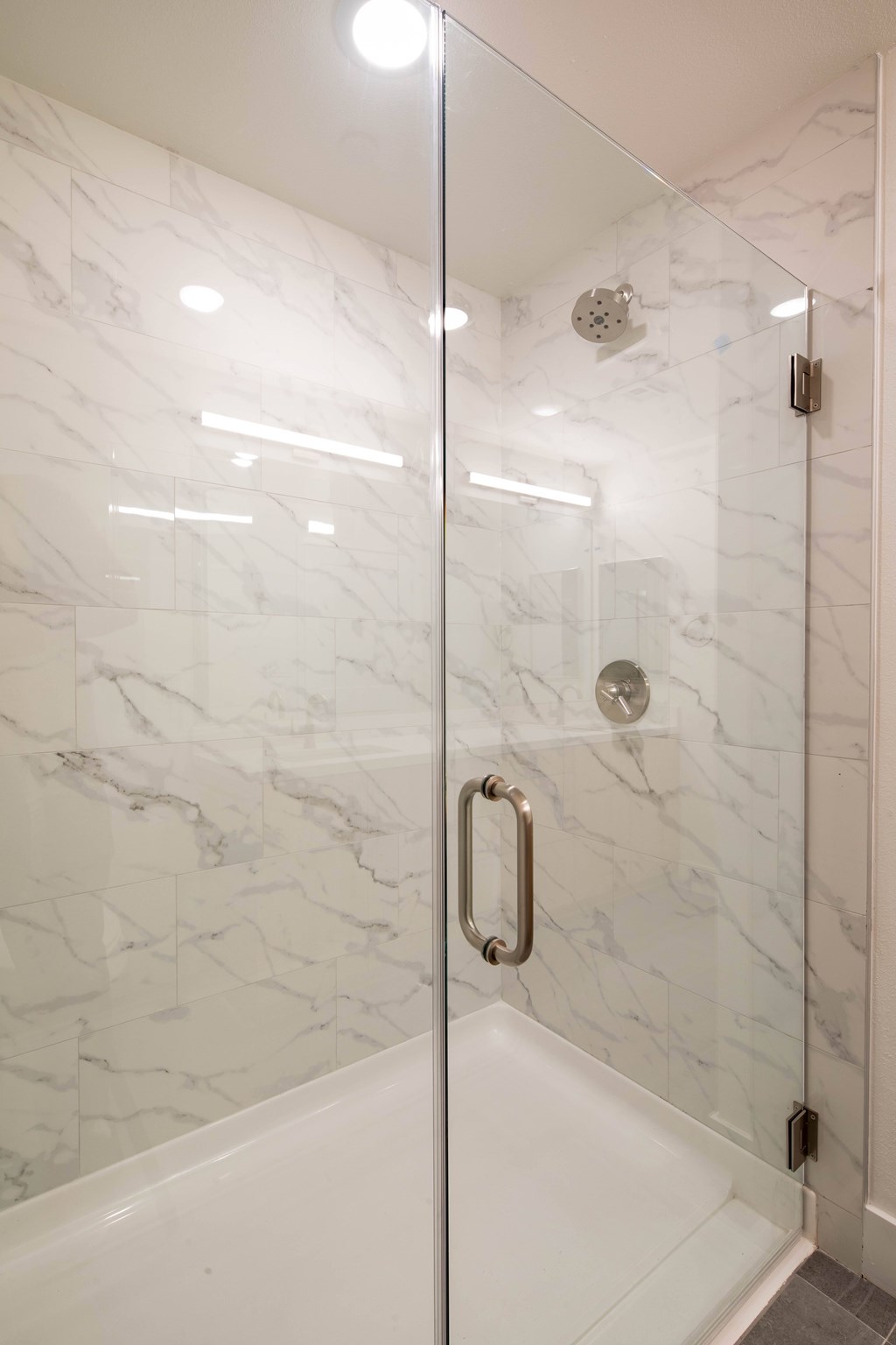 A white marble tiled shower with a glass door and a silver handle.