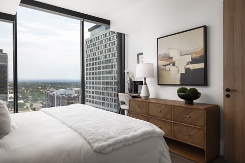 A bedroom with a large bed, a wooden dresser, and a large window overlooking a cityscape.