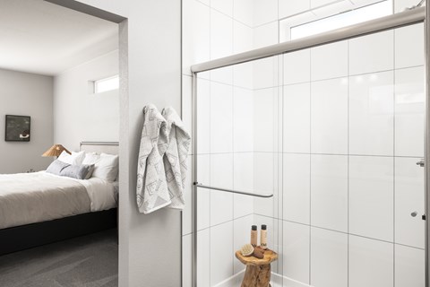 A bathroom with a white tiled shower and a bedroom with a bed and a nightstand.