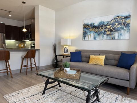 A modern living room with a grey couch, a glass coffee table, and a large abstract painting on the wall.