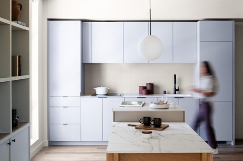 A woman is walking in a modern kitchen with white cabinets and a marble island.