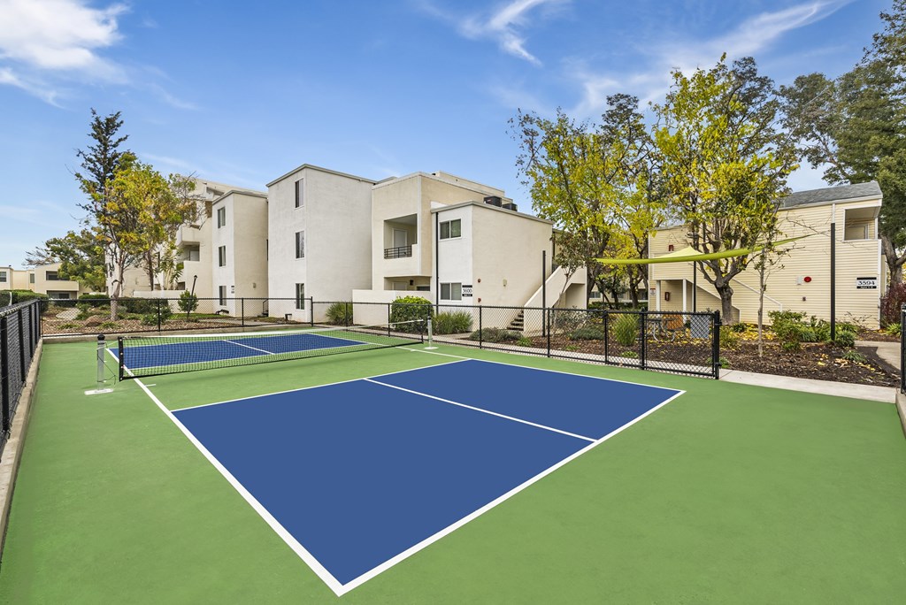 A tennis court with a blue surface and white lines is surrounded by a fence and trees.