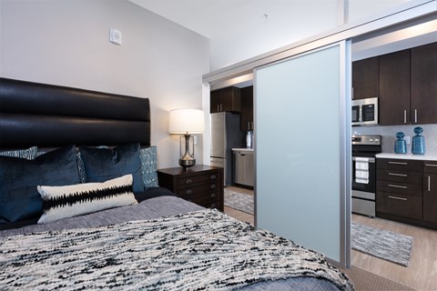 a bedroom with a bed and a nightstand with a lamp and a kitchen in the background