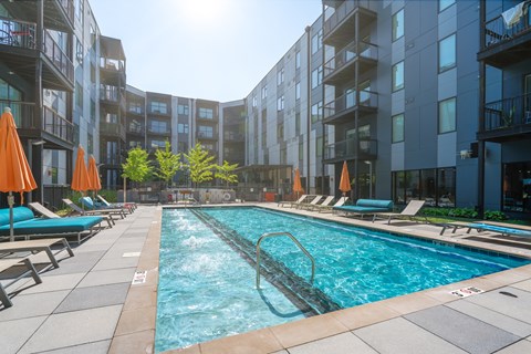A swimming pool surrounded by lounge chairs and umbrellas in front of apartment buildings.