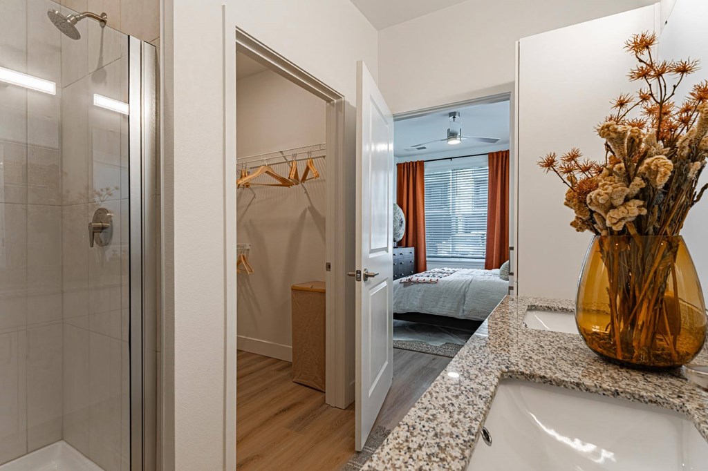 A bathroom with a walk-in shower, a bedroom, and a vase of dried flowers on the counter.