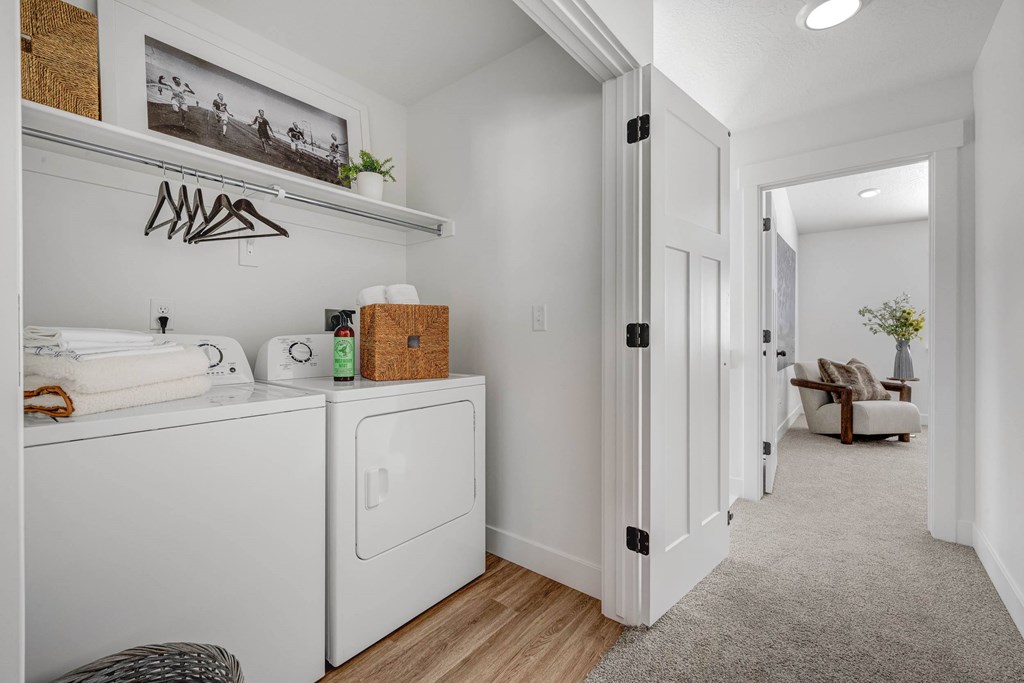 A laundry room with a washer and dryer, a shelf with clothes hangers, and a hallway leading to a living room.
