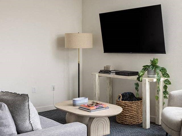 A living room with a grey couch, a white table, a lamp, a plant, and a television mounted on the wall.