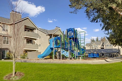 A playground with a blue slide and a green tree in front of apartment buildings.