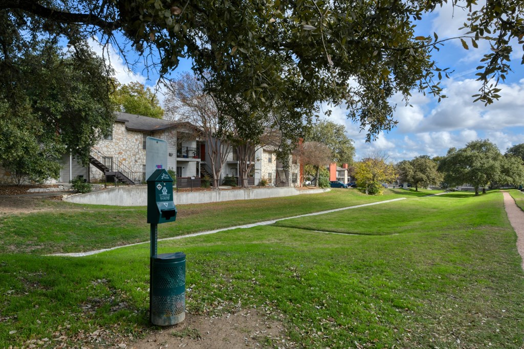 A green trash can is in the foreground of a grassy area with a path and houses in the background.