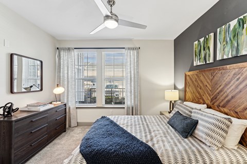 An interior view of a bedroom with a bed, a front end table with drawers, two large windows with curtains, a mirror, and a ceiling fan at Parkstone at Knightdale apartments for rent in Knightdale, NC.