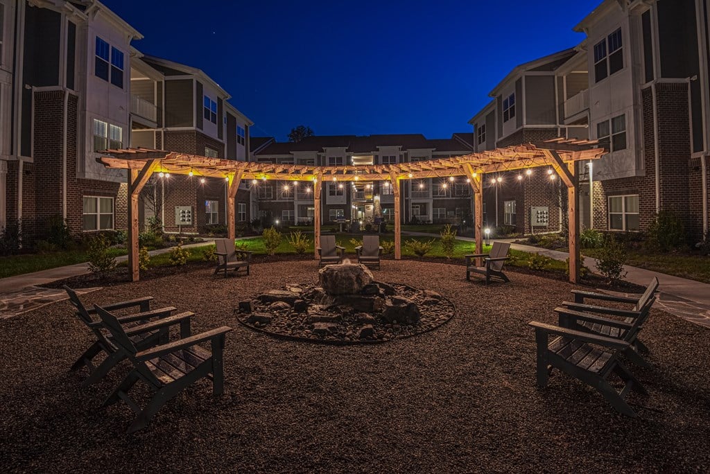 A fire pit is surrounded by chairs and a pergola with lights is above.