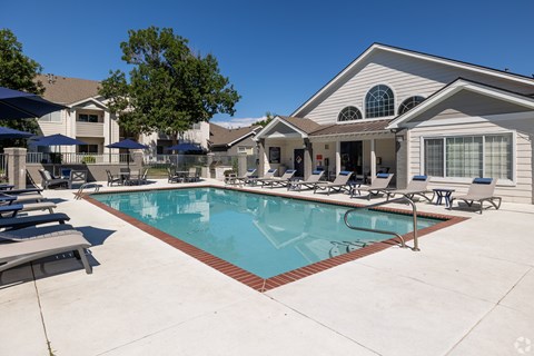 A swimming pool in front of a house with a patio and lounge chairs.
