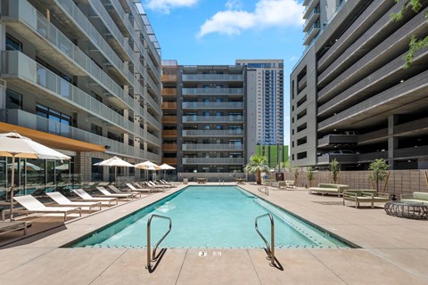 A large swimming pool surrounded by lounge chairs and umbrellas in front of apartment buildings.