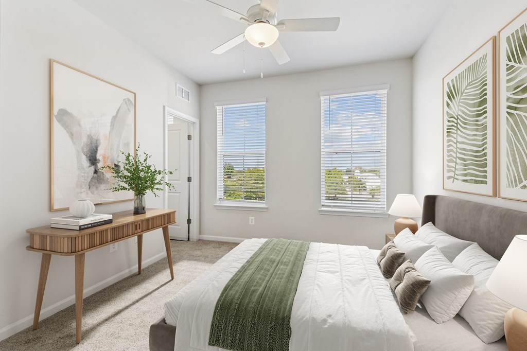 A bedroom with a bed, a table with a plant and a painting, and a window with blinds.