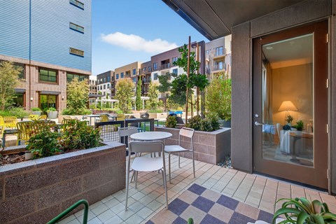 A patio with a table and chairs is surrounded by plants and buildings.