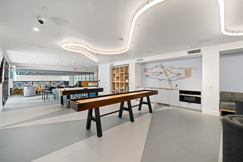 A long wooden table is in the middle of a white room with a white wall and a white framed picture.