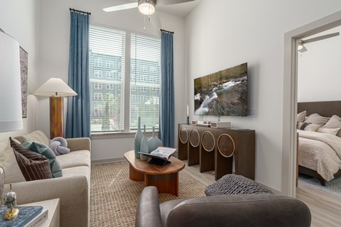 A well-lit, modern living room with a large window, a sofa, a chair, a coffee table, and a bed in the background.