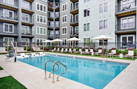 A swimming pool surrounded by lounge chairs and umbrellas in front of apartment buildings.