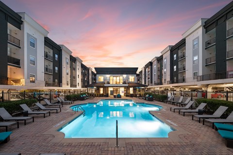 A large swimming pool surrounded by lounge chairs in front of apartment buildings.