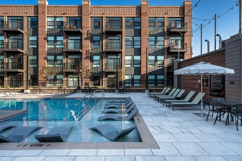 A large swimming pool in front of a brick building with lounge chairs and umbrellas.