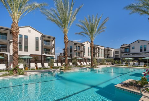A swimming pool surrounded by palm trees and lounge chairs in front of apartment buildings.