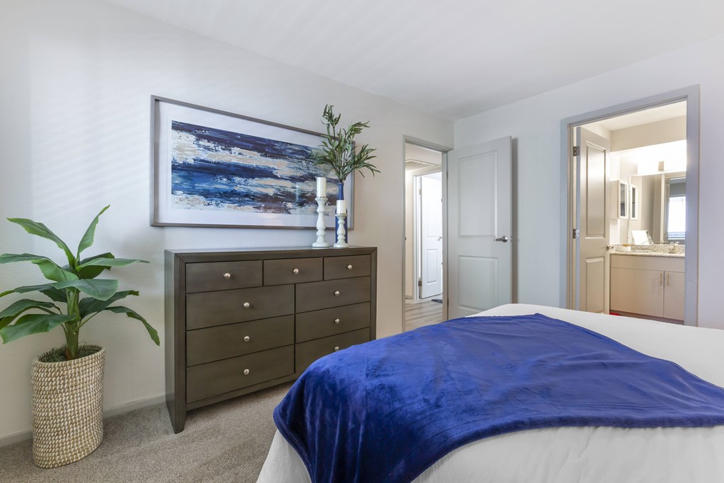 A bedroom with a bed covered in a blue blanket, a dresser, and a potted plant.