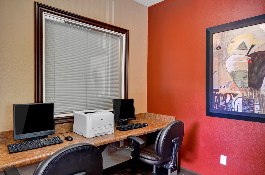 A desk with a computer monitor, keyboard, and printer in front of a window with blinds.