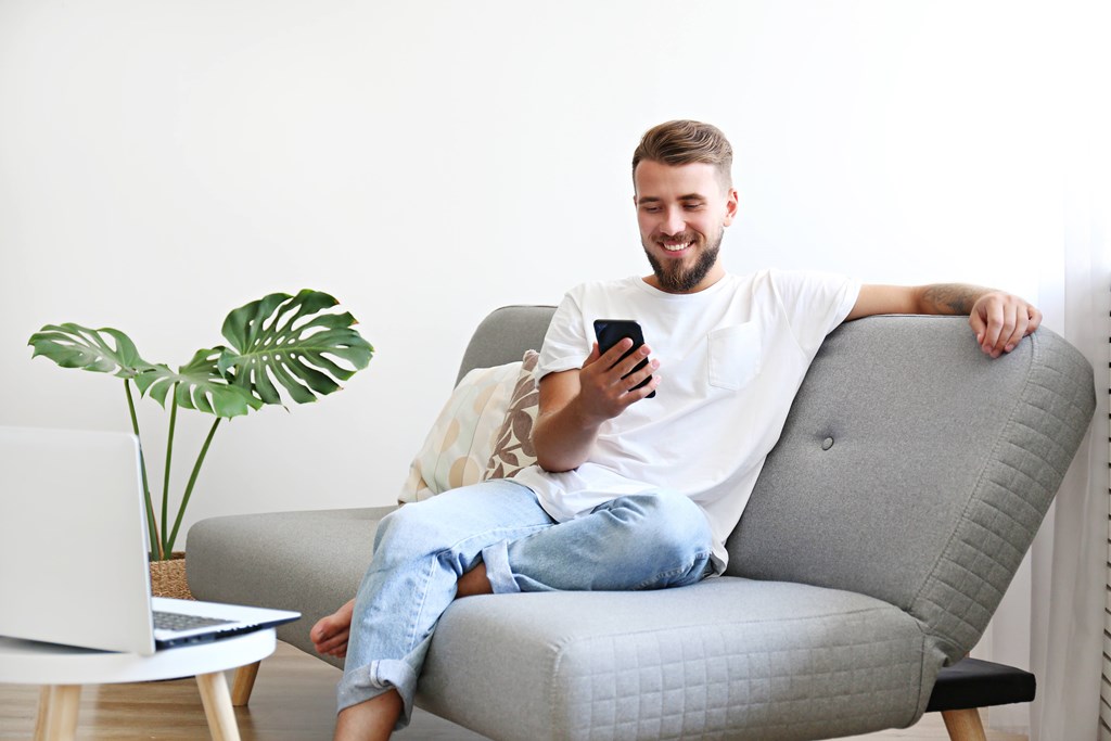 A man is sitting on a grey couch with a laptop and a phone in his hands.