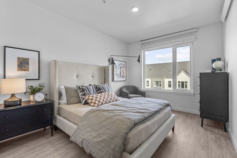 A bedroom with a large bed, a nightstand with a lamp, and a window with a view of a house.