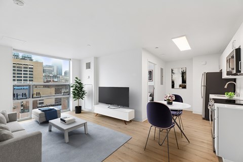 A modern living room with a grey sofa, a white coffee table, a black chair, a white TV stand, and a kitchen area with a sink and cabinets.