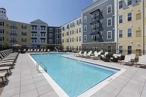 A large swimming pool surrounded by lounge chairs in front of apartment buildings.