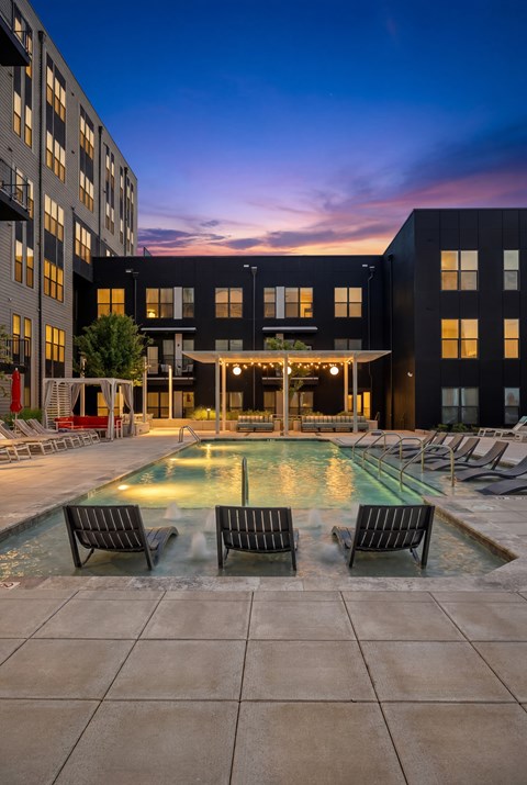 A pool surrounded by chairs and buildings at dusk.