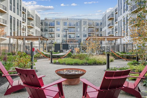 A fire pit surrounded by red chairs in a courtyard with buildings in the background.