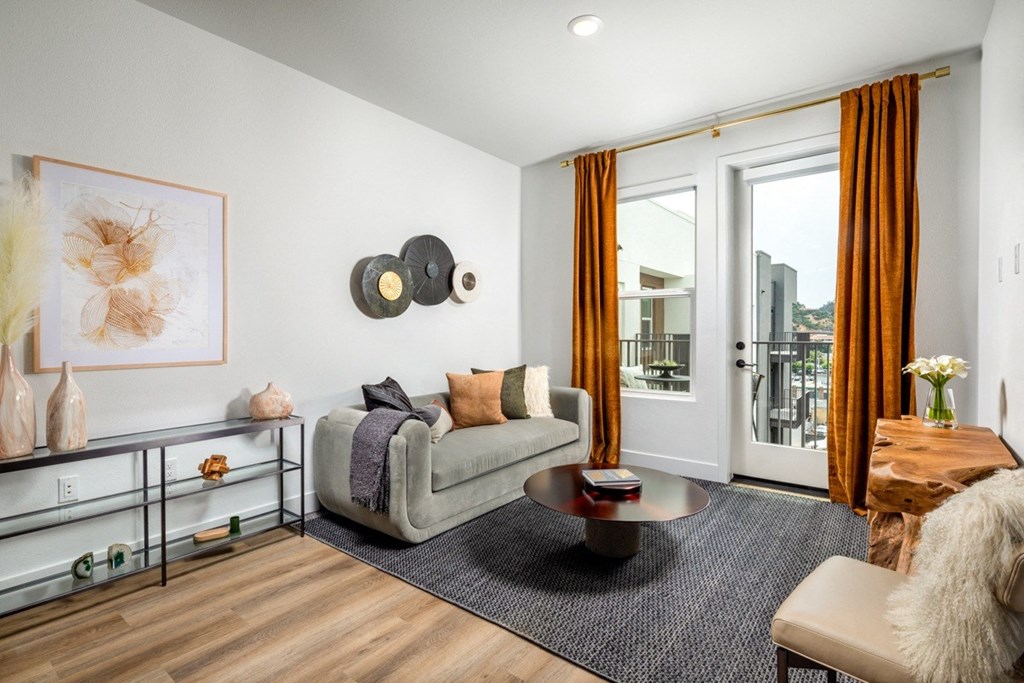 A living room with a grey couch, a coffee table, and a large window with orange curtains.