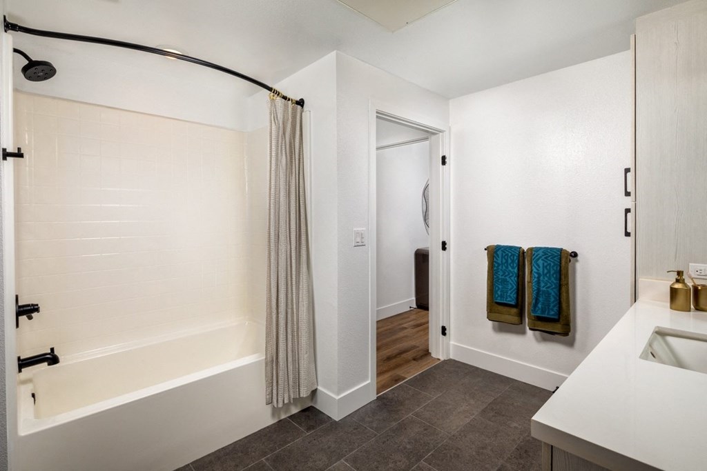 A bathroom with a white tub and sink, a shower with a curtain, and a doorway leading to another room.