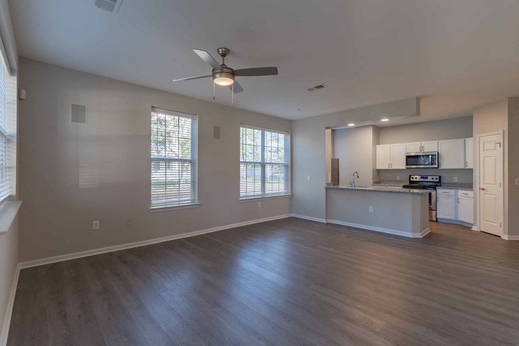 an empty living room with a ceiling fan and a kitchen in the background