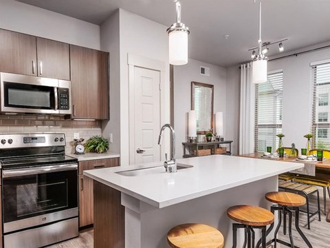 A modern kitchen with a stainless steel oven and microwave, a white countertop, and wooden bar stools.