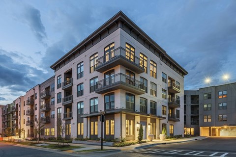 A modern apartment building with balconies and multiple windows is illuminated at dusk.