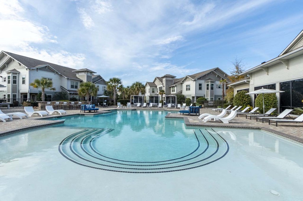 Two-Bedroom Apartments in Downtown Savannah, GA - Adara at Godley Station - Large Swimming Pool Surrounded by Sun Loungers, Cabanas, Umbrellas, Outdoor Couch, Landscaping, and a View of the Apartments.
