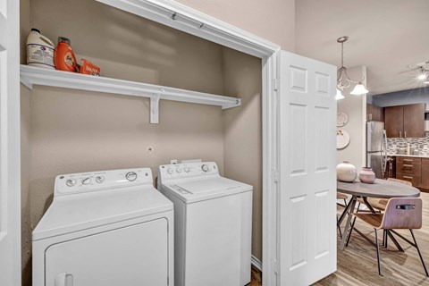 A laundry room with a washer and dryer, a shelf with some items, and a table with a chair.