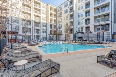 A large swimming pool surrounded by lounge chairs in front of apartment buildings.