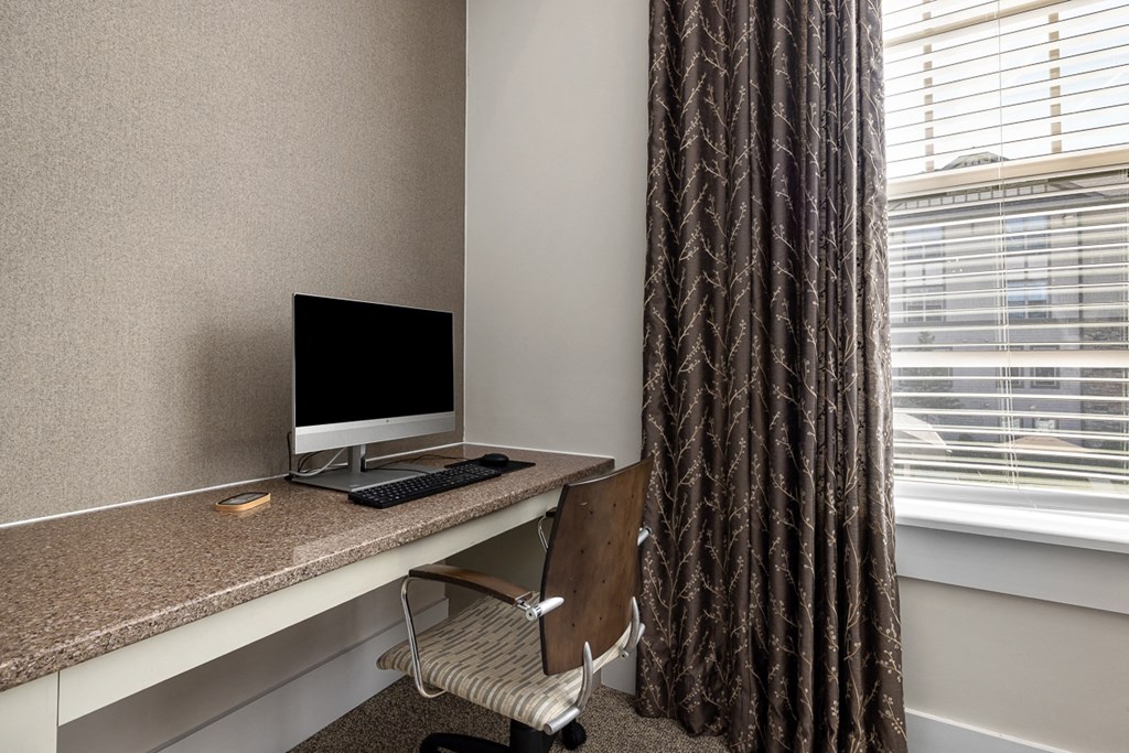 A computer desk with a monitor, keyboard, and chair in front of a window with blinds.