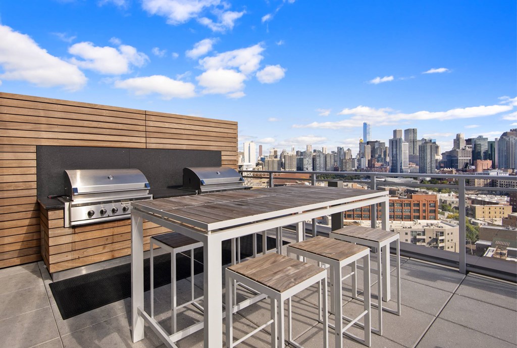 A rooftop patio with a grill and stools overlooking a city skyline.