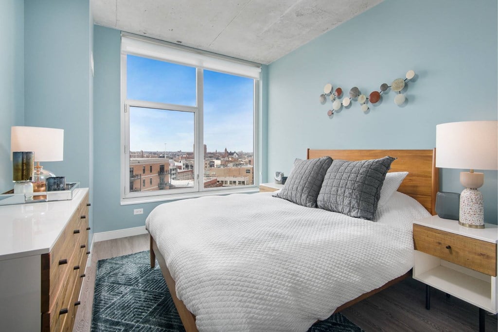 A bedroom with a large bed, a window with a view of the city, and a nightstand with a lamp.