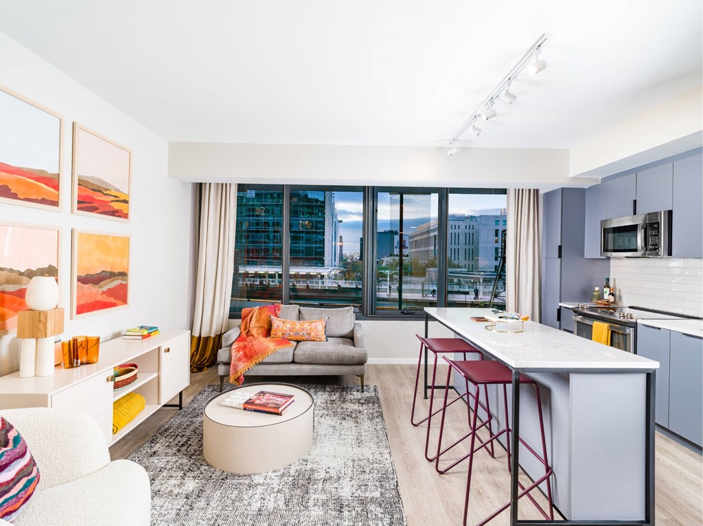 A modern living room with a grey sofa, a white coffee table, and a kitchen area with a white countertop and red bar stools.
