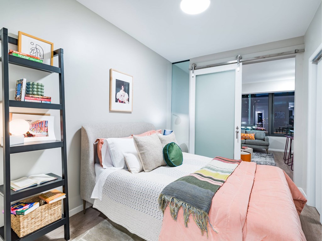 A bedroom with a bed, a bookshelf, and a window with a view of the city.