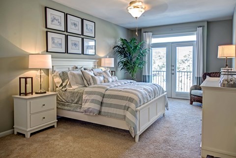 A bedroom with a large bed, a nightstand with a lamp, and a plant in the corner.