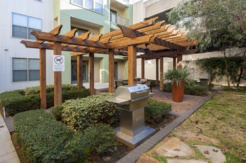 A wooden pergola is over a grill in a courtyard.