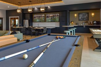 A pool table with a blue cloth and white cue balls is in the foreground of a room with a bar and seating area.